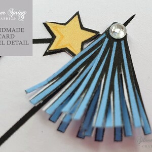 May include: A blue and black tassel with a white star and a rhinestone on top. The tassel is attached to a black stick. The text 'Handmade Card Tassel Detail' is in the background.