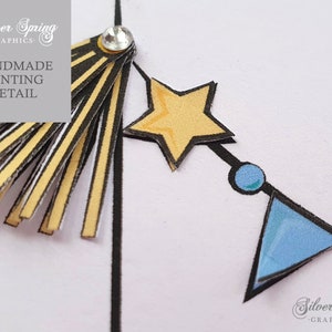 May include: A close-up of a handmade bunting detail with a yellow star, a blue circle, and a blue triangle. The bunting is made of paper and has a black outline. The bunting is attached to a string with a tassel at the top. The tassel is made of yellow and black paper strips. The text 'HANDMADE BUNTING DETAIL' is visible in the top left corner of the image.