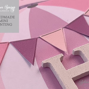 May include: Pink and white bunting with a wooden letter H. The bunting is made of felt and has a textured surface. The letter H is painted white and has a textured surface. The bunting is hanging from a string. The text "HANDMADE MINI BUNTING" is visible in the image.