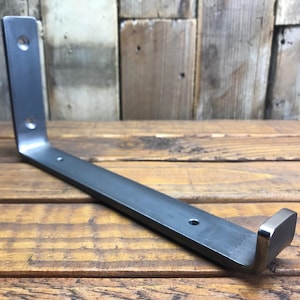 May include: A dark grey metal shelf bracket with a right-angle design. The bracket has three pre-drilled holes for mounting and a small, squared-off end. The metal has a matte finish and appears to be made of steel.