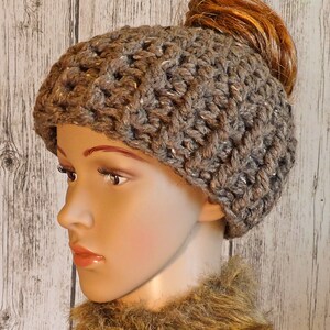 Op de afbeelding: Een grijze gehaakte beanie met een gat bovenop voor een paardenstaart. De beanie wordt gedragen door een mannequin met een bruine nep bont sjaal.
