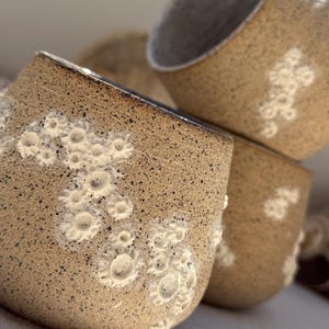 May include: A stack of four handmade ceramic cups. The cups are a speckled tan color with white floral designs and a dark brown rim. The interior of the cups is a light gray color. The cups are stacked at an angle.
