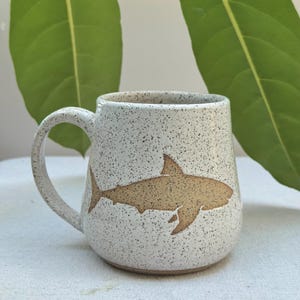 Könnte beinhalten: Gesprenkelter Keramikbecher mit einem Haifisch-Design. Der Becher hat eine weiße Basis mit dunklen Sprenkeln und einer braunen Haifisch-Silhouette. Er hat einen gebogenen Griff und einen abgerundeten Körper. Der Becher ist für heiße oder kalte Getränke geeignet.