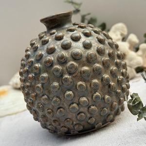 May include: A small, round ceramic vase with a textured surface of raised, rounded bumps. The vase has a dark, mottled finish with shades of brown, green, and bronze. The opening is small and cylindrical. Decorative home decor.