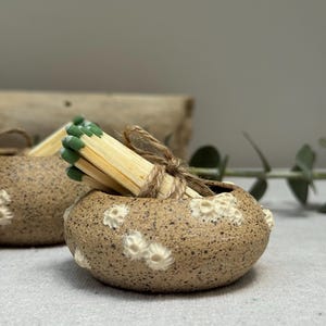 May include: A small, speckled ceramic match holder with white floral accents. A bundle of wooden matches with green tips is tied with twine and placed inside. The holder is a light brown color.