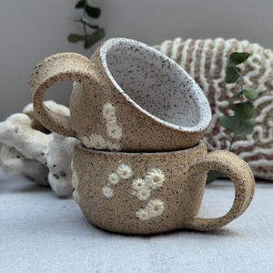 Może przedstawiać: Dwa ręcznie robione ceramiczne kubki ułożone jeden na drugim. Kubki są w kolorze nakrapianego brązu z białymi akcentami i białym wnętrzem. Każdy kubek ma duży uchwyt.