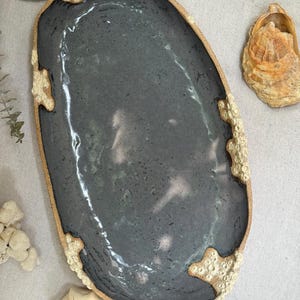 May include: An oval ceramic tray with a smooth, dark charcoal-colored glazed center and a rough, textured, beige-colored rim that features raised, coral-like organic details along the edges, creating a contrast between the dark basin and light border.