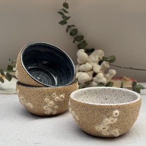 Pode incluir: Três pequenas tigelas de cerâmica feitas à mão. As tigelas têm um exterior bege salpicado com detalhes brancos texturizados. O interior de uma tigela é azul escuro, enquanto outra é branca salpicada. As tigelas estão sobre uma superfície branca.