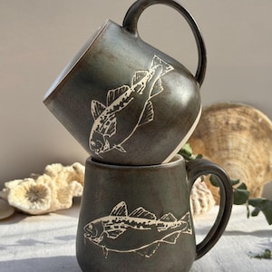 May include: Two dark gray ceramic mugs stacked, each featuring a white etched fish design. The mugs have large handles and a rustic, textured appearance. The background includes natural elements, suggesting a coastal or nature-inspired theme.