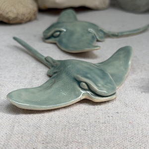 May include: Two ceramic stingray sculptures with a glossy, sage green glaze. Each piece features a flat, wide body with extended wing-like fins and a long, slender tail. The surface of the clay has a smooth, reflective finish with subtle color variations.