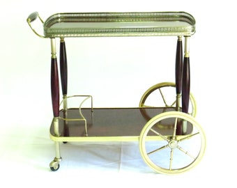 Puede incluir: Un carro de servicio vintage de latón y madera con una tapa de espejo y dos ruedas. El carro tiene una base de madera oscura y detalles de metal dorado.