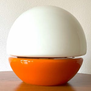 Könnte beinhalten: Eine weiße und orangefarbene Keramik-Tischlampe mit einem runden, kuppelförmigen Schirm.