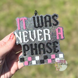 Puede incluir: Adorno gris brillante con el texto "IT WAS NEVER A PHASE" en negro, blanco y rosa. El adorno tiene un patrón de cuadros en la parte inferior y un pequeño lazo para colgar. El logotipo de Freshie Library es visible.