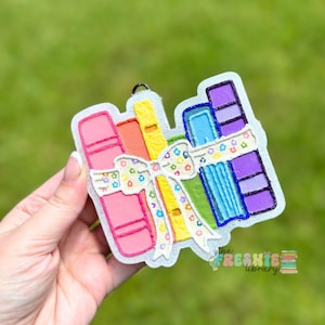 May include: A vibrant charm depicting a stack of books in pink, orange, yellow, blue, and purple, tied with a white bow adorned with flowers. The charm has a silver outline and a small loop for hanging. The text "the freshie library" is visible.