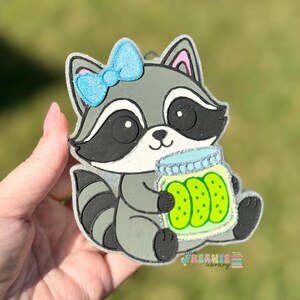 May include: A gray raccoon-shaped ornament with a blue bow and a jar of pickles. The raccoon has black and white markings and is holding a jar of green pickles. The ornament has glitter accents and the text "FRESHIE library".