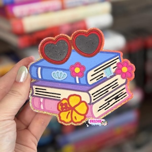 May include: A decorative item featuring a stack of three books in blue, white, and pink, topped with red heart-shaped sunglasses. The design includes a yellow hibiscus flower, a small pink flower, and a blue shell icon. The text Freshie Library is visible.