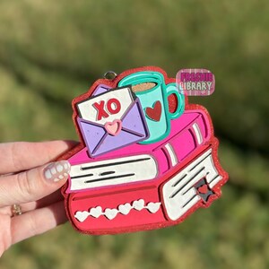 Könnte beinhalten: Ein Valentinstag-Ornament mit einem Bücherstapel, einer Tasse mit einem Herz und einem Umschlag mit "XO". Das Ornament ist rot, rosa, lila und türkis, mit einem glitzernden Finish. Der Text "Freshie Library" ist ebenfalls sichtbar.