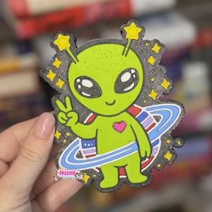 May include: A lime green alien figure with large black eyes, a pink heart on its chest, and a peace sign hand gesture. The alien is surrounded by yellow stars and a blue planet ring. The text FRESHIE reads in small letters at the bottom left.
