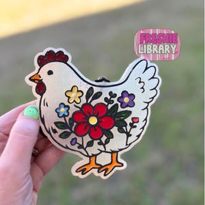 May include: A white, glittery chicken-shaped ornament with a black outline. The chicken is decorated with colorful flowers in red, yellow, blue, and purple. The ornament has orange feet and a red comb. The text "FRESHIE LIBRARY" is in a pink and white striped box.