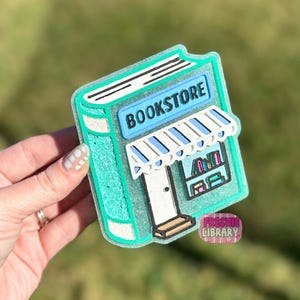 May include: A teal-colored bookstore-shaped charm with the word "BOOKSTORE" in a blue rectangle. The charm features a white awning with blue stripes, a white door, and a small shelf with books. The charm is held by a hand.