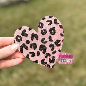 May include: A pink heart-shaped decoration with a glittery leopard print design. The black spots are scattered across the pink surface. The decoration has the text "FRESHIE LIBRARY" on a pink and white sticker.