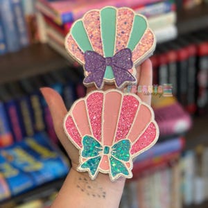 May include: Two seashell-shaped decorative items. One is pink and the other is teal, both with glitter accents and a bow. The items have a silver outline and are held in a hand. The text "the fresbie library" is visible.