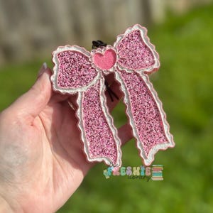 May include: A pink glitter bow with a white scalloped trim and a small pink heart in the center. The bow has two long, flowing ribbons and a black string for hanging. The bow is a decorative item.