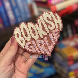 May include: A heart-shaped accessory with the words "BOOKISH GIRLIE" in white and pink glitter. The accessory is rose gold and has a sparkly finish. The brand name "the FRESHIE" is at the bottom.