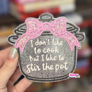 May include: A silver glittery pot-shaped air freshener featuring a pink glittery bow at the top. The text reads, I don't like to cook but I like to stir the pot. The design is finished with a small logo in the bottom right corner.