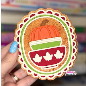 May include: A circular scalloped ornament featuring a glittery copper background. The center displays a stacked design of an orange pumpkin sitting atop a light green bowl, which rests inside a dark red bowl decorated with three white leaf icons.