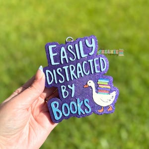 May include: A purple glittery ornament with the text "EASILY DISTRACTED BY BOOKS". It features a white goose carrying a stack of colorful books. The ornament has a small silver loop for hanging.
