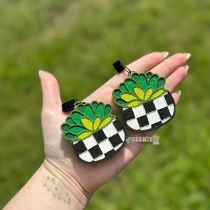 May include: Two decorative succulent-shaped air fresheners. Each features a green and yellow plant design in a black and white checkered pot. They have a black clip for hanging. The word "FRESHIE" is visible.