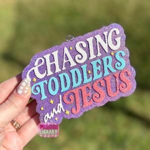 Può includere: Un ornamento viola glitterato con le parole "CHASING TODDLERS and JESUS" in bianco, turchese e rosa. L'ornamento ha un bordo decorativo e piccoli accenti di stelle gialle. Le parole sono in un carattere stravagante.