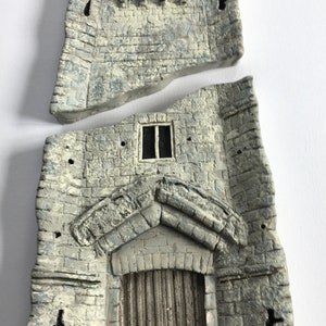 Può includere: Un muro di castello in pietra grigia con una porta e una finestra. Il muro è rotto in due pezzi. La porta ha una porta in legno e una cornice in pietra ad arco. La finestra è rettangolare e ha due vetri.