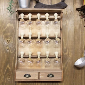 Puede incluir: Un estante de especias de madera para colgar en la pared con dos cajones y 15 tarros de vidrio con tapones de corcho. El estante est&aacute; hecho de madera marr&oacute;n claro y tiene un aspecto r&uacute;stico.