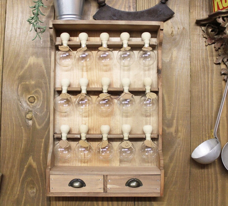 Puede incluir: Un estante de especias de madera para colgar en la pared con dos cajones y 15 tarros de vidrio con tapones de corcho. El estante est&aacute; hecho de madera marr&oacute;n claro y tiene un aspecto r&uacute;stico.