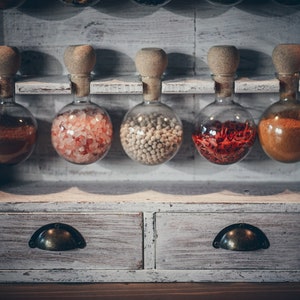 May include: A white wooden spice rack with six glass jars filled with different spices. The jars have cork tops and are arranged in a row on the top shelf. The rack has two drawers with metal handles.