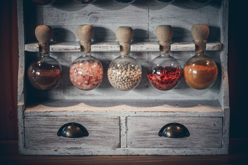 May include: A white wooden spice rack with six glass jars filled with different spices. The jars have cork tops and are arranged in a row on the top shelf. The rack has two drawers with metal handles.