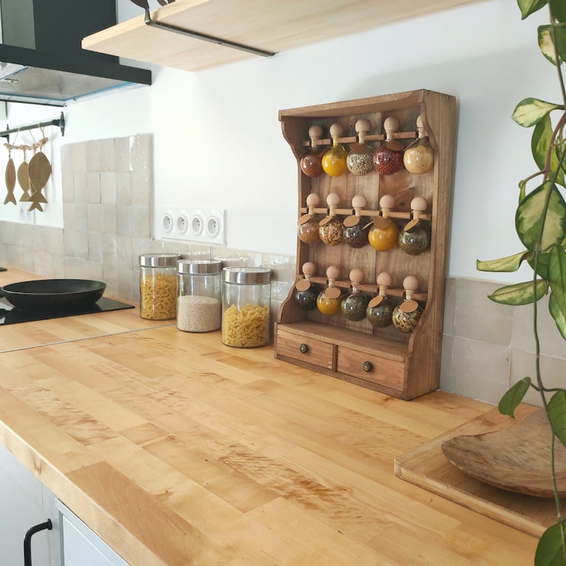 Puede incluir: Estante de especias de madera con dos estantes y frascos de vidrio llenos de especias. El estante est&aacute; montado en una pared sobre una encimera de madera clara.