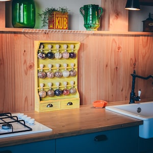May include: A yellow wooden spice rack with glass jars filled with spices. The rack has two drawers and is mounted on a wooden wall. The rack is in a kitchen setting with a stovetop and a sink.