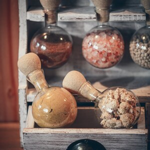 May include: A white wooden shelf with six glass jars with cork stoppers. The jars contain various spices and dried flowers. The jars are arranged in two rows of three. The shelf is on a wooden surface.