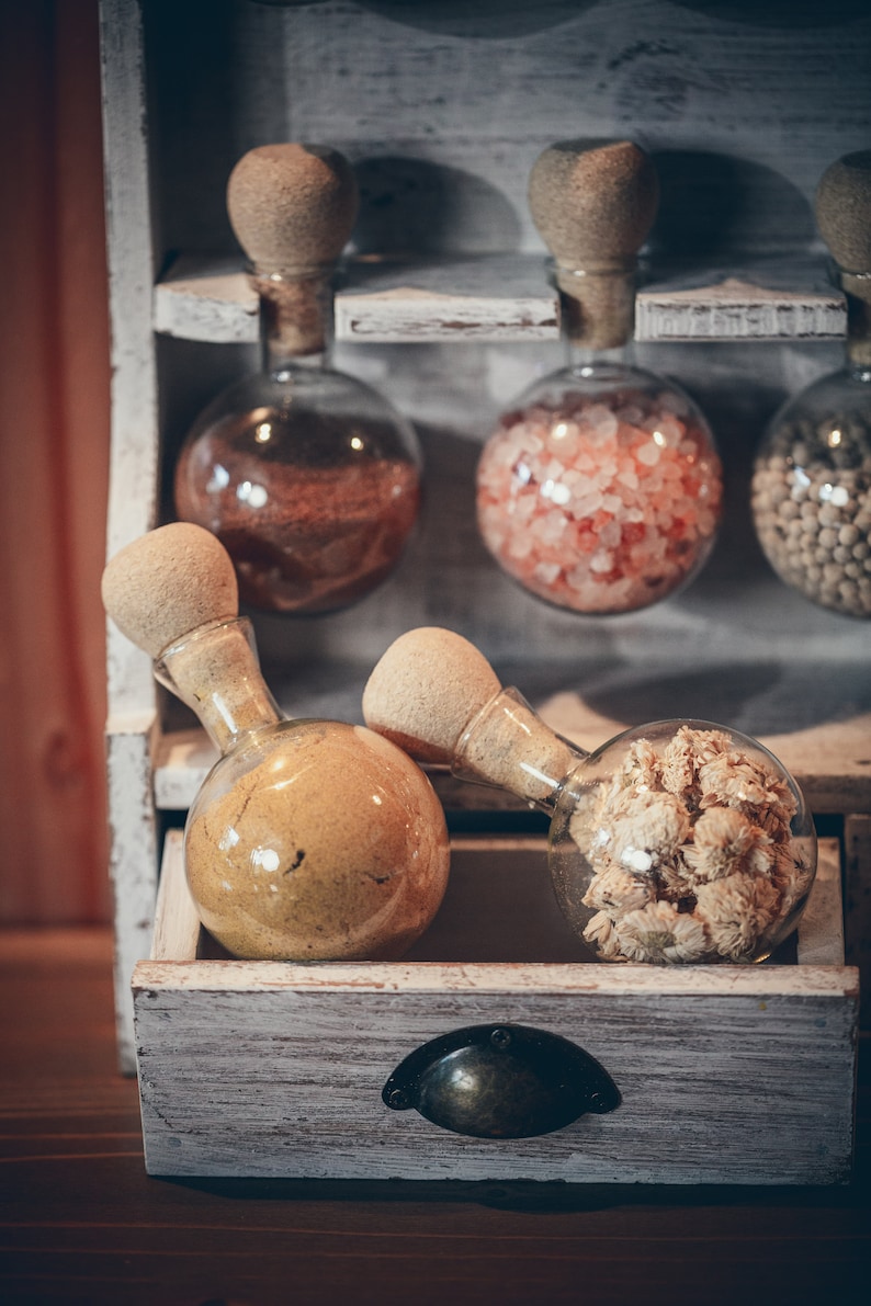 May include: A white wooden shelf with six glass jars with cork stoppers. The jars contain various spices and dried flowers. The jars are arranged in two rows of three. The shelf is on a wooden surface.