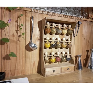 Puede incluir: Estante de especias de madera con dos cajones y 18 frascos de vidrio llenos de diferentes especias. Los frascos tienen tapas de corcho y est&aacute;n dispuestos en tres filas de seis. El estante est&aacute; montado en una pared de madera.