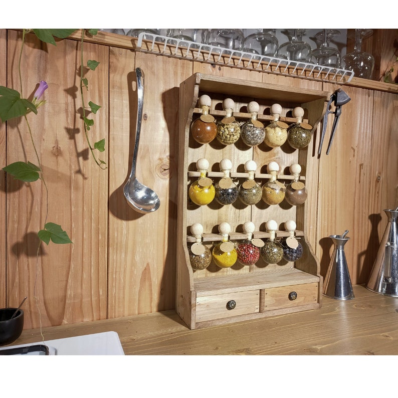 Puede incluir: Estante de especias de madera con dos cajones y 18 frascos de vidrio llenos de diferentes especias. Los frascos tienen tapas de corcho y est&aacute;n dispuestos en tres filas de seis. El estante est&aacute; montado en una pared de madera.