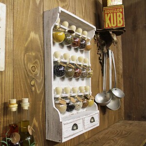 May include: White wooden spice rack with glass jars filled with various spices. The rack has two drawers and is mounted on a wooden wall.