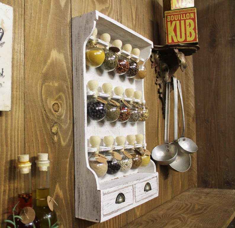 May include: White wooden spice rack with glass jars filled with various spices. The rack has two drawers and is mounted on a wooden wall.