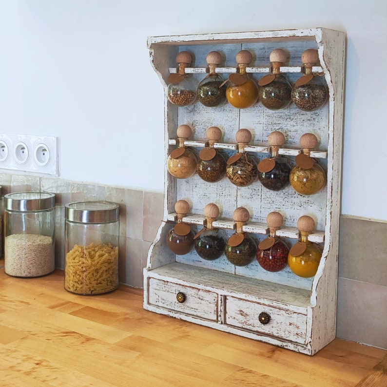 May include: White painted wooden spice rack with two drawers and 12 glass jars with cork tops. The jars are filled with various spices and herbs. The rack is mounted on a wall.
