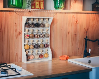Può includere: Un porta spezie in legno bianco con 24 barattoli di vetro trasparente pieni di diverse spezie. Il porta spezie è montato su una parete in legno con una finitura in legno chiaro. Il porta spezie si trova in un ambiente da cucina con un piano cottura in acciaio inox e un piano di lavoro bianco.