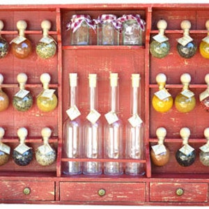 May include: A red wooden spice rack with three drawers and multiple glass jars filled with spices. The jars are hung on hooks and have wooden tops. There are also several clear glass bottles on the shelf.