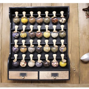 May include: A black wooden spice rack with two drawers, holding 36 small glass jars with various spices and herbs. The jars have wooden lids with a knob on top. The rack is mounted on a wooden wall.